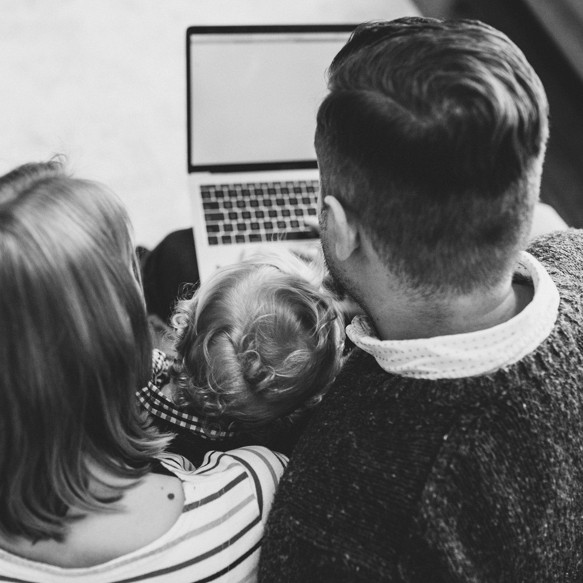 Two people and child viewed from behind, gathered around a laptop. Black and white image, suggesting family bonding or shared activity.